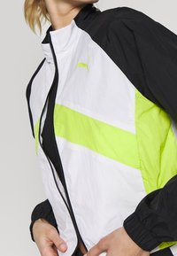 Puma vindjacka med svarta, vita och fluorescerande gula paneler; lätt, med dragkedja, elastiska muddar och en sportig design.
