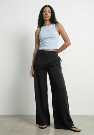 Kvinde med krøllet hår iført en lyseblå ærmeløs crop top, sorte bukser med vide ben og blonder, samt sorte klipklapper, stående mod en hvid væg.