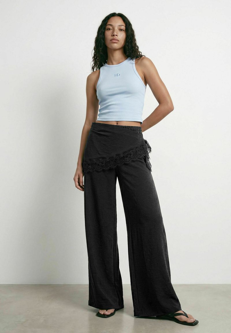 Femme aux cheveux bouclés portant un crop top sans manches bleu clair, un pantalon noir à jambes larges avec un détail en dentelle, et des tongs noires, debout contre un mur blanc.