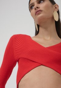 Frau in einem roten gerippten, verkürzten Pullover mit Ausschnitt, großen strukturierten goldenen Ohrringen, die nach oben blickt, vor einem schlichten Hintergrund.