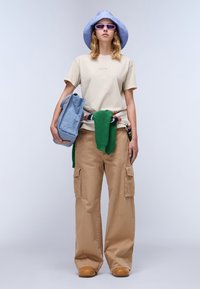 Beige t-shirt met de tekst "NAPA", groene gebreide omslag, khaki cargobroek, blauwe tas en een paarse strooien hoed, gedragen met bruine klompen.