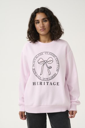 Femme aux cheveux bouclés et foncés portant un sweat-shirt rose clair avec un motif de nœud et les mots Heritage, Classic Cotton, Vintage Nostalgia.