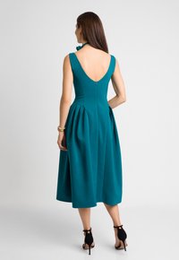 Kobieta w turkusowej, bezrękawnikowej sukience midi z głębokim dekoltem na plecach i plisowanym dołem, stoi na czarnych szpilkach na jednolitym tle.