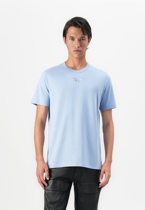 Ung mand iført en lyseblå T-shirt med korte ærmer og et lille sølvlogo på brystet samt sorte læder cargo-bukser, stående mod en hvid baggrund.