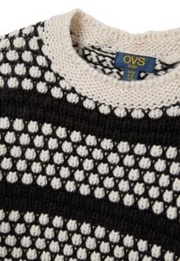 Gestrickter Pullover mit einem Muster aus schwarzen und cremefarbenen Rauten, mit einem gerippten Ausschnitt und weicher Textur. Etikett zeigt Größe 4-5 Jahre an.