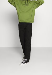 Grön huvtröja med framficka, kombinerad med svarta cargobyxor med sidofickor och svarta sneakers med vita sulor.