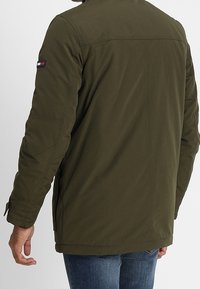 Olivgrön jacka med en slät textur, med ståkrage, synliga sömmar och en logotypapplikation på vänster ärm.