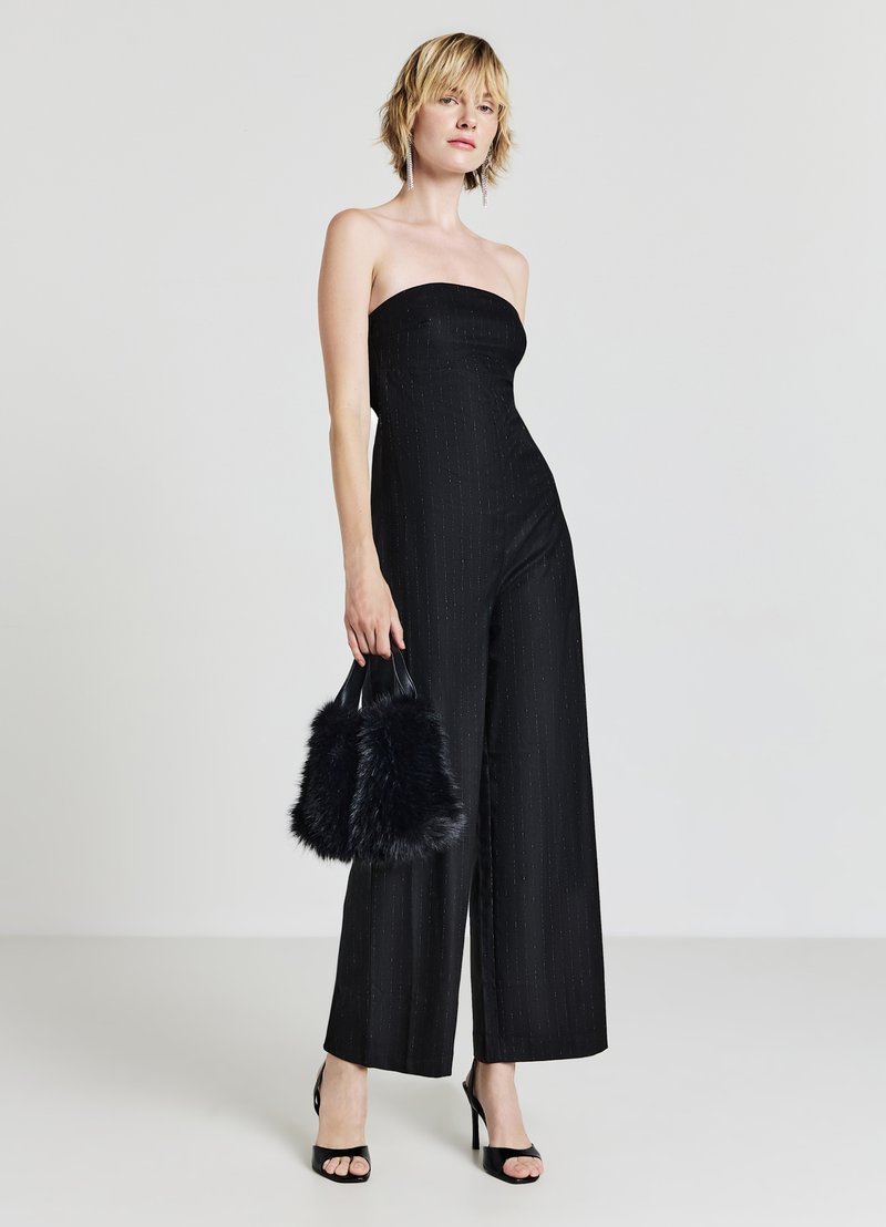 Schwarzer trägerloser Jumpsuit mit weitem Bein und Streifenmuster. Kombiniert mit einer schwarzen Handtasche aus Kunstfell und schwarzen hochhackigen Sandalen.