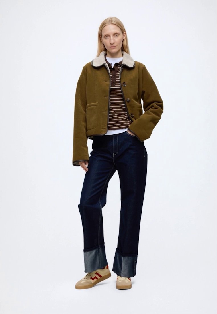 Femme debout portant une veste vert olive avec un col en fourrure, un pull marron rayé, un jean foncé retroussé et des baskets beige avec des bandes rouges.