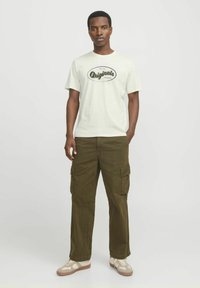 Lichtgekleurde katoenen t-shirt met de tekst "Originals"; olijfgroene cargobroek met zijzakken; tan en witte sneakers.