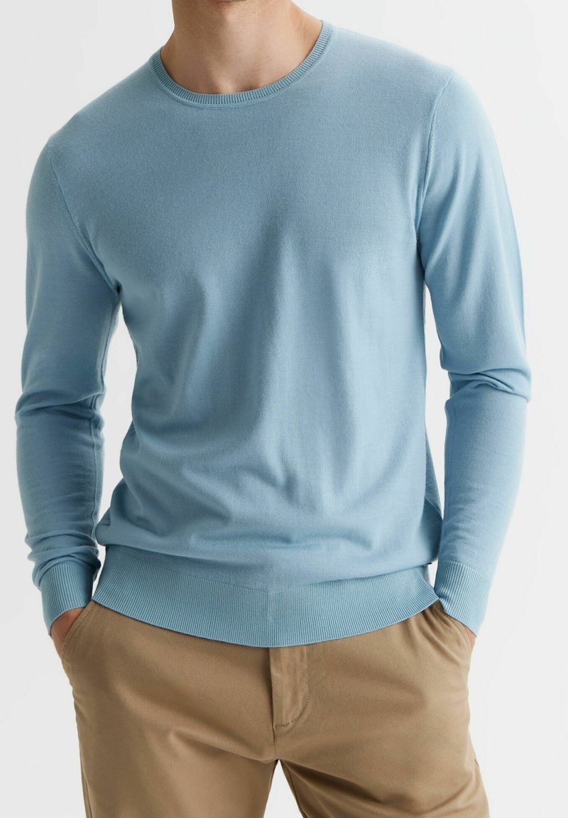 Homme portant un pull léger bleu clair à col rond et un pantalon beige, les mains dans les poches, torse visible sur fond blanc.