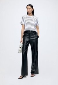 Woman wearing a white and black striped t-shirt, black wide-leg leather pants, black sandals, and holding a small white handbag.