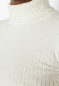 Gros plan sur une personne portant un pull à col roulé blanc côtelé, montrant la texture et le motif vertical en tricot.