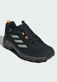 Zapatos exteriores negros con parte superior de tela texturizada, rayas grises reflectantes, acentos naranjas y una suela de goma resistente.
