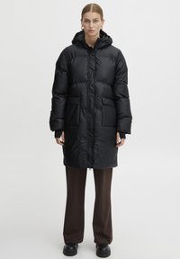 Lungo cappotto imbottito nero con cappuccio, dotato di zip frontale, due grandi tasche e una texture morbida e lucida. Indossato sopra dei pantaloni marroni.