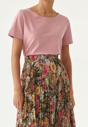 Frau trägt ein schlichtes rosa Kurzarm-Top, das in einen hoch taillierten, plissierten Rock mit roten, gelben und weißen Blumen gesteckt ist.