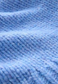 Měkká pletená látka světle modré barvy s výrazným vzorem a viditelnými stehy. Materiál vypadá nadýchaně a hřejivě.