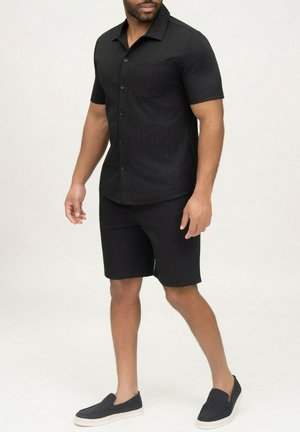 Homme portant une chemise noire à manches courtes boutonnée et un short noir assorti, avec des chaussures noires sans lacets.