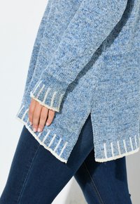 Ljust blå stickad tröja med vit fransad nederkant och sidofickor, med ett strukturerat mönster. Parat med mörk denim.