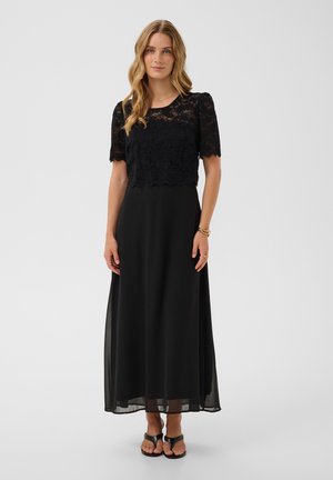 Femme aux longs cheveux blonds portant un top noir en dentelle à manches courtes et une longue jupe noire, debout devant un fond uni.