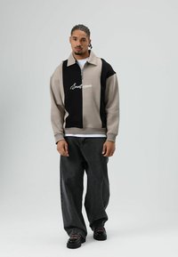 Beige en zwarte sweater met een ritskraag, voorzien van een logo. Gecombineerd met losse zwarte jeans en grove zwarte schoenen.