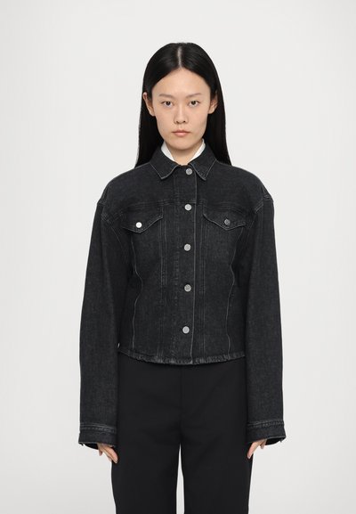 Una donna con capelli neri lunghi indossa una giacca di denim nera con bottoni argentati e pantaloni neri, in piedi contro uno sfondo grigio uniforme.