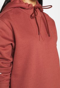 Mikina barvy rezavě hnědé s texturovanou látkou, kapucí na stažení a žebrovanými manžetami. Design zahrnuje švy jako detail.