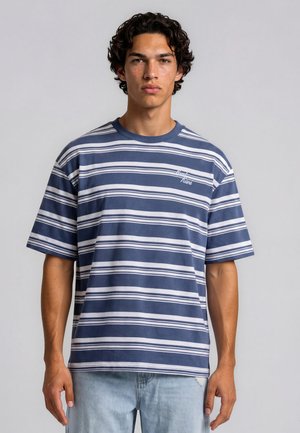 Jeune homme aux cheveux noirs bouclés et taches de rousseur portant une chemise rayée bleue et blanche à manches courtes et un jean bleu clair sur un fond uni.