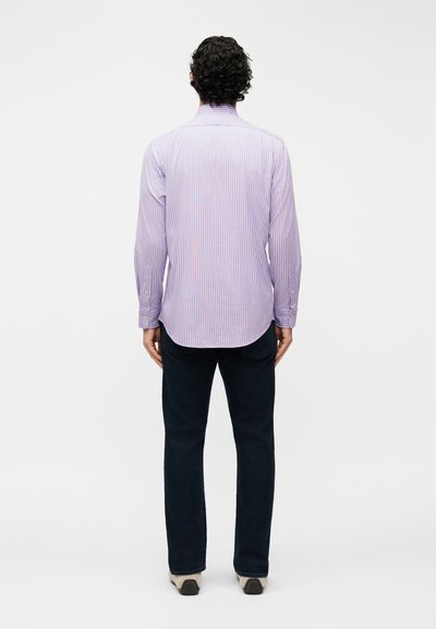 Άνδρας με σγουρά μαλλιά που φοράει ένα ανοιχτό μωβ ριγέ πουκάμισο μακρυμάνικο, σκούρα παντελόνια και μπεζ παπούτσια, στέκεται γυρισμένος με την πλάτη σε λευκό φόντο.