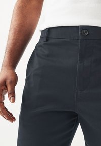 Pantaloni di cotone blu scuro con una texture liscia, dotati di chiusura con bottone e tasche laterali, mostrati brevemente accanto a una mano.