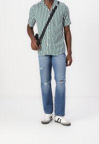 Muž s krátkým rukávem zeleno-bíle pruhované vertikálně, roztrhané modré džíny, bílé tenisky a s černou crossbody taškou.