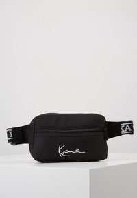 Czarna torba na talię z zamkiem błyskawicznym, zdobiona białymi detalami sygnatury i kontrastowym czarno-białym paskiem.