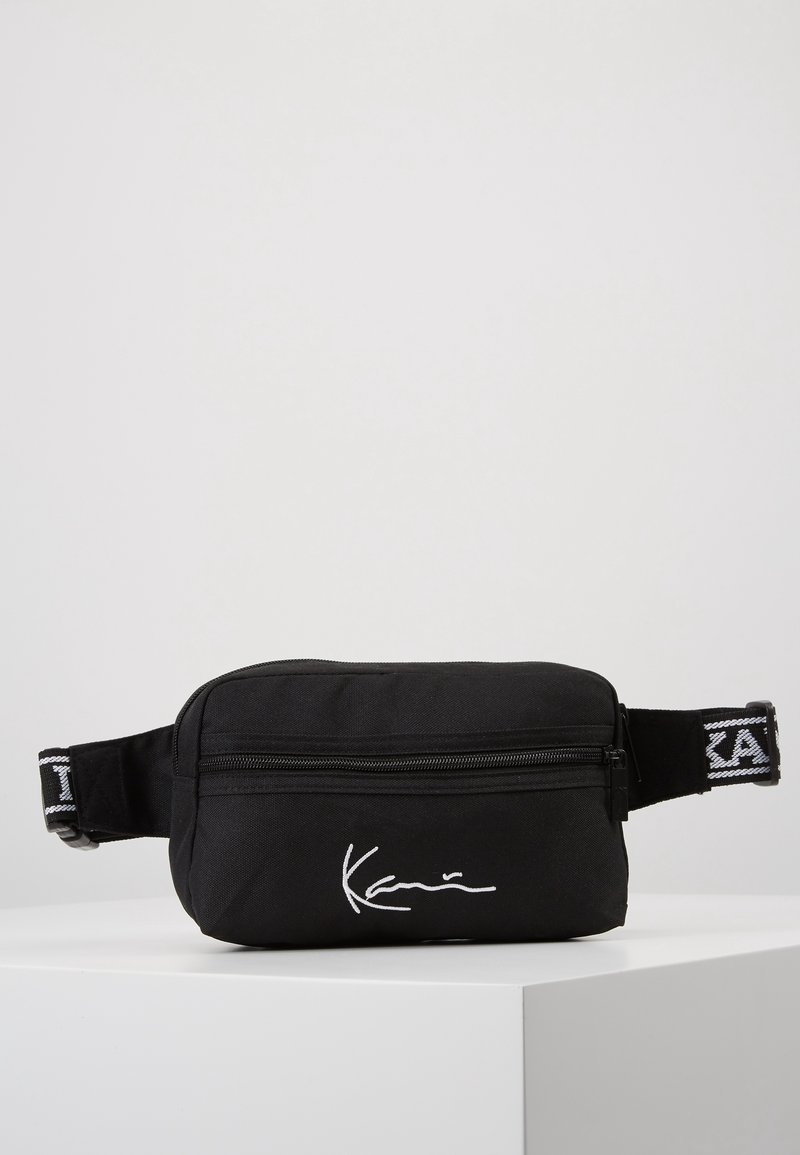 Czarna torba na talię z zamkiem błyskawicznym, zdobiona białymi detalami sygnatury i kontrastowym czarno-białym paskiem.