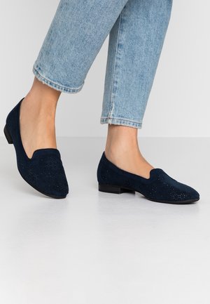 Kvinde iført lyseblå cropped jeans og navyblå perforerede ruskindslædersko med lave hæle, der står på hvidt gulv.