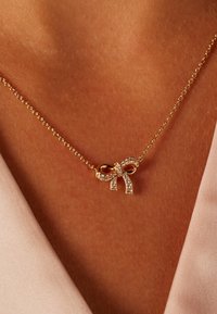 Collier en or avec un pendentif en forme de nœud orné de strass transparents. La chaîne présente un design délicat et se porte sur une peau nue en arrière-plan.