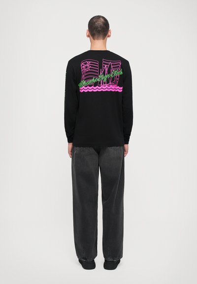 Chemise noire à manches longues avec un design graphique rose et vert comportant des palmiers, des bateaux et des vagues. Associée à un pantalon gris ample.