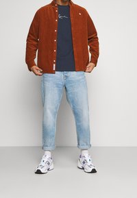 Camisa de pana color óxido, camiseta navy, jeans azul claro remangados y zapatillas blancas con acentos morados.