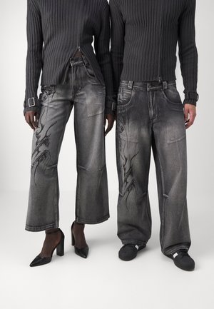 Deux personnes portant des pulls à zip côtelés noirs et des jeans gris délavés à jambes larges avec des motifs abstraits noirs, l'une en talons, l'autre en baskets.