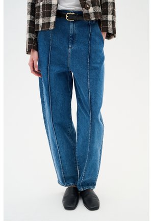 Blauwe jeans met hoge taille en voorste naden, zwarte riem met gouden gesp, bruin-wit geruit jasje, zwarte schoenen met vierkante neus.