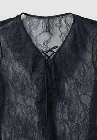 Blusa nera in pizzo con scollatura a V, parte anteriore arricciata e dettagli con lacci al collo. Materiale traslucido con motivi intricati.