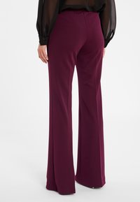 Pantalons bordeaux à jambe large avec une texture lisse, dotés d'une taille haute et de détails de coutures subtils, présentés de dos sur fond blanc.