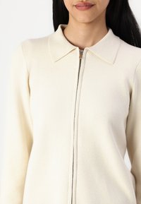 Cardigan crema con zip e colletto a punta, caratterizzato da una texture morbida e un design minimalista, che presenta una chiusura con zip argentata.