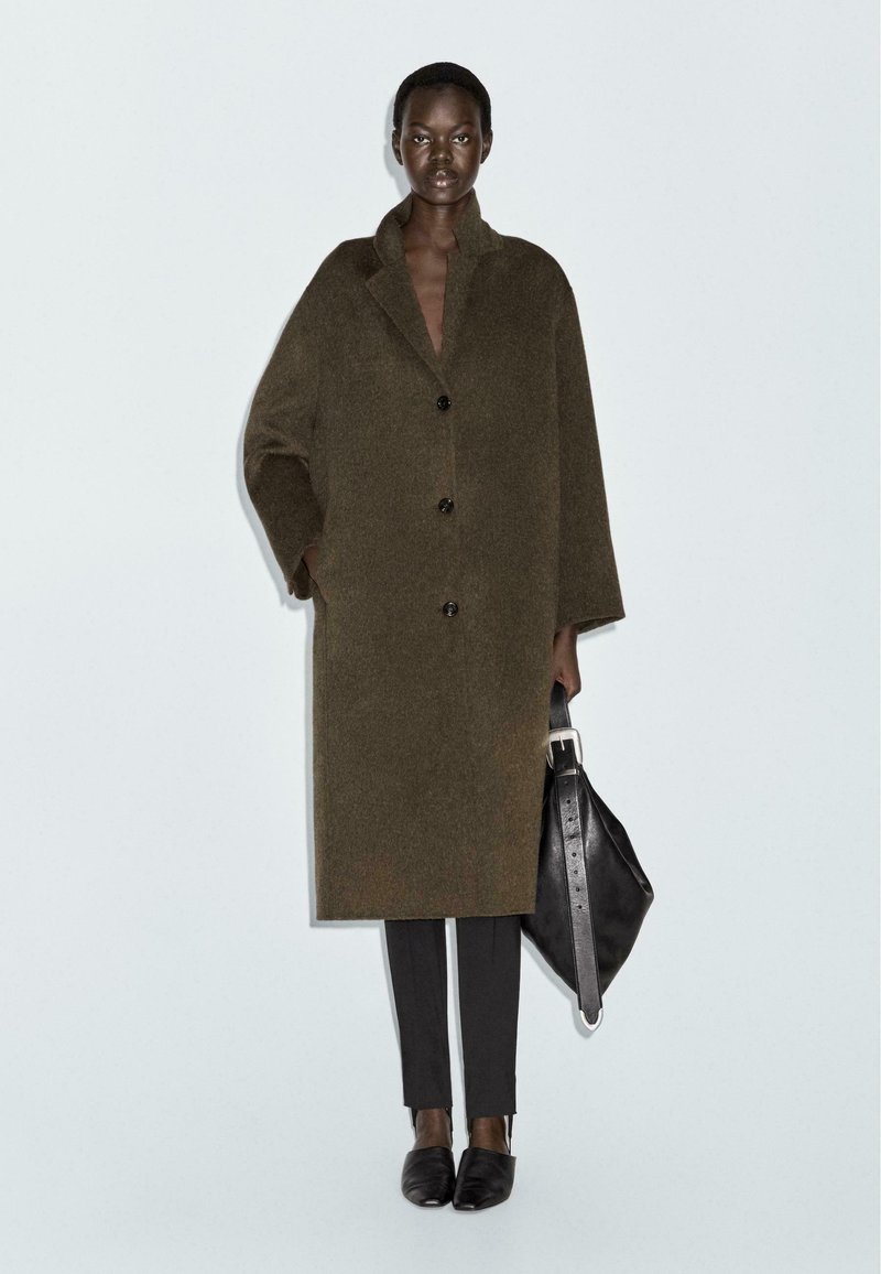 Cappotto in lana verde oliva con vestibilità ampia, chiusura a cinque bottoni, grandi revers, indossato sopra pantaloni neri e con una borsa in pelle nera.
