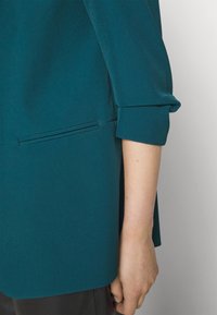 Blazer bleu sarcelle avec une texture lisse, doté de manches retroussées, de détails de poches latérales et d'une coupe décontractée.