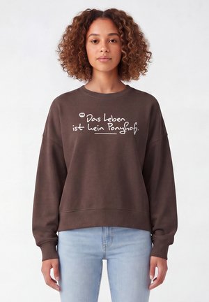 Frau mit lockigem Haar trägt braunen Pullover mit deutschem Text "Das Leben ist kein Ponyhof" und hellblauen Jeans, stehend vor weißem Hintergrund.