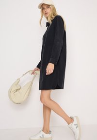 Schwarzes Cordhemdkleid mit langen Ärmeln, seitlichen Schlitzen und einem Kragen. Kombiniert mit weißen Sneakern und einer beigen Schultertasche.