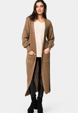 Kvinde iført en lang brun strikket cardigan over en beige top, sorte læderbukser og sorte platformstøvler, stående med hænderne i lommerne.