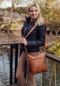 Braune Leder-Crossbody-Tasche mit glatter Textur, Reißverschlussfächern und verstellbarem Tragegurt. Lässig kombiniert mit einer schwarzen Daunenjacke und beigen Leggings.