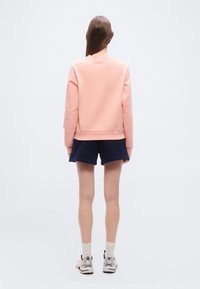 Άτομο που στέκεται με την πλάτη, φοράει ανοιχτό ροζ φούτερ, σκούρο μπλε σορτς, λευκές κάλτσες και λευκά αθλητικά παπούτσια σε απλό φόντο.