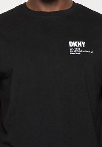 Černé tričko s bílým nápisem na hrudi „DKNY založeno 1989, oficiální uniforma New Yorku.“